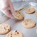 A hand in gloves puts a chocolate chip cookie on a Vollrath aluminum perforated sheet pan.