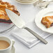 A knife being used to cut a pie on a plate with a piece of pie and a cup of coffee on it.