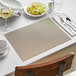 A table set with Front of the House tan woven vinyl placemats, silverware, and wine glasses.