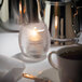 A Hollowick clear glass tealight candle holder on a table next to a teapot and coffee cup.