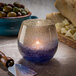 A Hollowick Laredo blue bubble glass votive with a lit candle on a table with a bowl of olives and a knife.