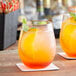 Two Visions clear plastic stemless wine glasses filled with orange liquid and garnished with a slice of orange and mint on a table.