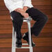A man wearing Uncommon Chef black chef pants sitting on a stool.