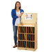 A woman standing next to a Jonti-Craft wood classroom sign-in station shelf with slots for folders.