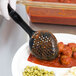 A gloved hand uses a black Vollrath Spoodle to serve food onto a white plate with red sauce and peas.