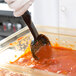 A gloved hand uses a Vollrath High Heat Perforated Oval Nylon Spoodle to scoop sauce into a container.