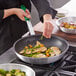 A person using a Choice 14" aluminum non-stick fry pan with a green silicone handle to cook food.