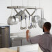 A man in a stainless steel kitchen with a Regency ceiling-mounted pot rack holding ladles and pots.