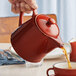 A person pouring tea into a red Acopa Keystone stoneware teapot.