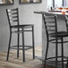A Lancaster Table & Seating black metal ladder back bar stool with a black wood seat next to a table.