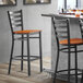 A Lancaster Table & Seating black ladder back bar stool with a cherry wood seat next to a table.