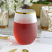 A Visions clear plastic stemless wine glass with a gold rim filled with red liquid on a table.