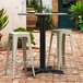 A Lancaster Table & Seating black outdoor table base with white stools on a brick patio.