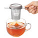 A hand holding an OXO stainless steel tea infuser over a cup of tea.