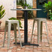 A black Lancaster Table and Seating outdoor table base with white stools on a brick patio.