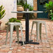 A Lancaster Table & Seating Excalibur black outdoor table base with stools on a brick patio.