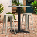 A Lancaster Table & Seating black outdoor table base with a bar height column on a brick patio with white bar stools.