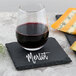 A glass of red wine on a slate coaster with the word "merlot" next to cheese.