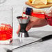 A person using an OXO stainless steel double jigger to pour red liquid.