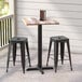 A Lancaster Table & Seating Excalibur counter height table with a textured wooden top and cross base plate on a porch with two black stools.
