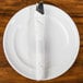 A fork and knife wrapped in a white customizable paper napkin band on a plate.