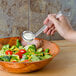 A hand holding a Cambro clear plastic ladle pouring dressing into a bowl of salad.