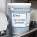 A bucket of Noble Chemical Dry It Plus rinse aid on a counter in a professional kitchen.