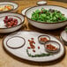 A GET glazed cream melamine bowl with brown trim on a table with bowls of fruit, nuts, and green salad.