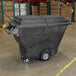 A large black Rubbermaid tilt truck with a hinged dome lid sits in a warehouse.
