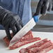 A hand in a black glove using a Dexter-Russell Santoku knife to cut raw meat.