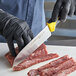 A person in a black glove cutting raw meat on a counter with a Dexter-Russell yellow Santoku knife.