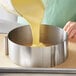 A person using an Ateco stainless steel adjustable cake ring to pour yellow liquid into a cake pan.