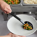 A hand holding a Carlisle black perforated salad bar spoon over a bowl of vegetables.