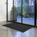 A black Lavex Needle Rib indoor entrance mat unrolled in front of a glass door.