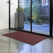 A burgundy Lavex Olefin entrance mat on the floor in front of a glass door.