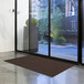 A brown Lavex Needle Rib entrance mat unrolled on the floor in front of a glass door.