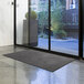 A rolled charcoal Lavex Olefin entrance mat on the floor in front of a glass door.