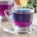 A person's hand holding a lemon over a glass cup of Wild Hibiscus Blue-Tee Butterfly Pea Flower herbal tea with purple liquid.