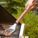 A hand with a rosewood-handled stainless steel turner cooking meat on a grill.