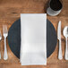 A wooden plate with silverware and a white Milan satin band cloth napkin on it.