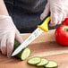 A person wearing a plastic glove with a yellow and black Mercer Culinary Millennia utility knife cutting a cucumber.