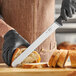 A person wearing black gloves uses a Schraf 12" serrated slicing knife to cut bread on a cutting board.