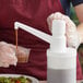 A gloved hand using a Tablecraft stationary condiment pump to pour brown liquid into a white container on a counter.