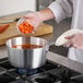 A person in a white coat stirring a red liquid in a silver Choice Tapered Aluminum Sauce Pan on a professional kitchen counter.