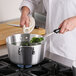 A person in a white coat cooking broccoli in a Choice aluminum sauce pan on a stove.