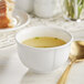 A white Acopa porcelain bouillon cup filled with soup and a spoon on a table with bread.
