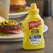 A close up of a French's Classic Yellow Mustard squeeze bottle next to a burger.