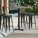 A Lancaster Table & Seating bar height table with a textured black metal finish and cross base plate on a patio with chairs and stools.