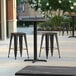 A Lancaster Table and Seating square counter height table with a cross base on a patio with black stools.