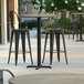 A Lancaster Table & Seating bar height table with a textured metal finish and cross base plate on an outdoor patio with black metal chairs.